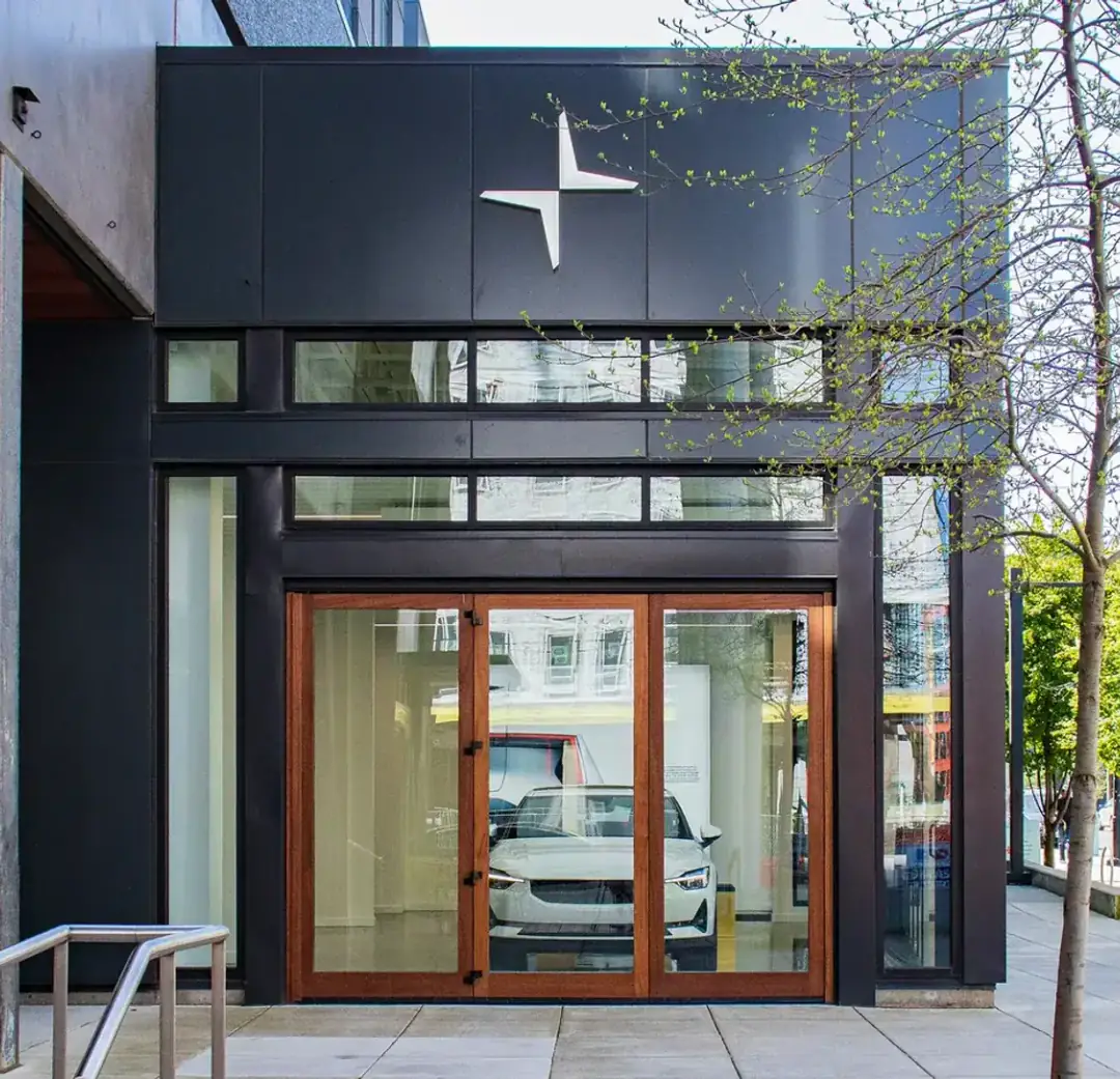 The dark-clad frontage of a car showroom at street level, lit interior and timber-framed entry doors behind full-height glazing