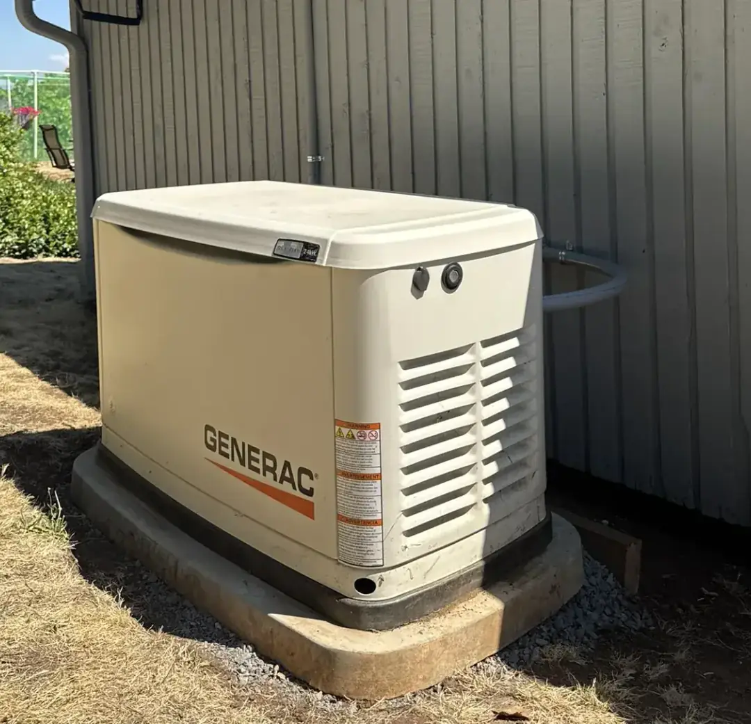 Generac standby generator on a concrete pad beside a board-and-batten farmhouse, fuel line run into the wall behind it